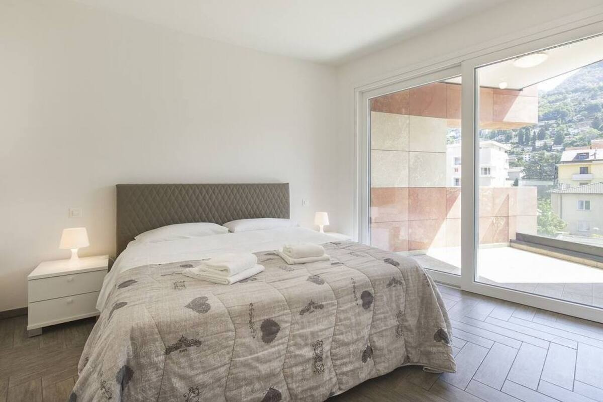 A bedroom with a bed, nightstands, and a large window with a view of buildings and hills.