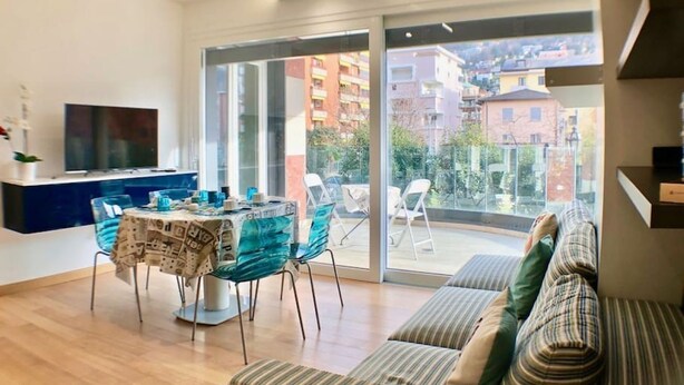 A modern living room with a sofa, a dining table with chairs, a TV, and a sliding glass door leading to a balcony. - Roggia Apartments (Lugano)
