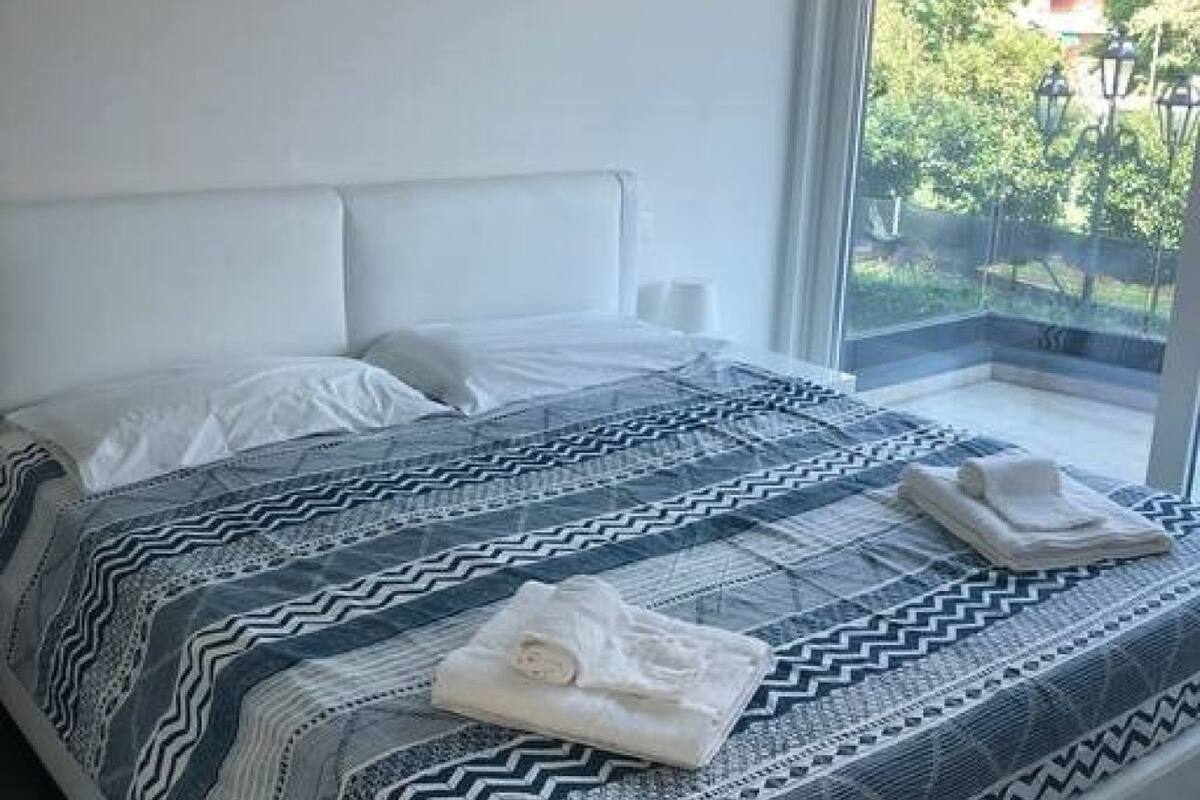 A hotel room with a bed, white headboard, and a view of greenery outside.
