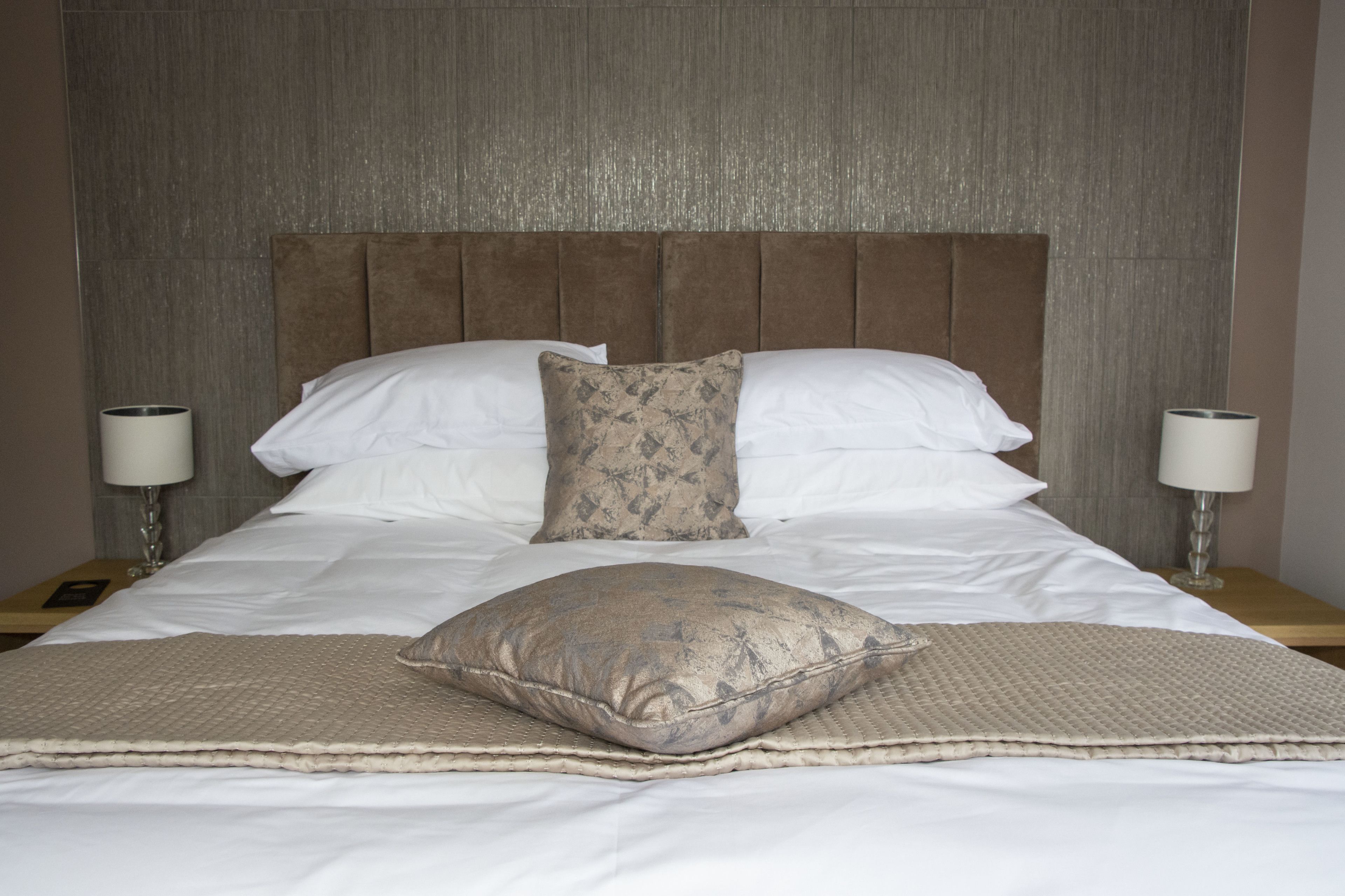 Una cama bien hecha con cabecera, dos lámparas de noche y un cojín decorativo.