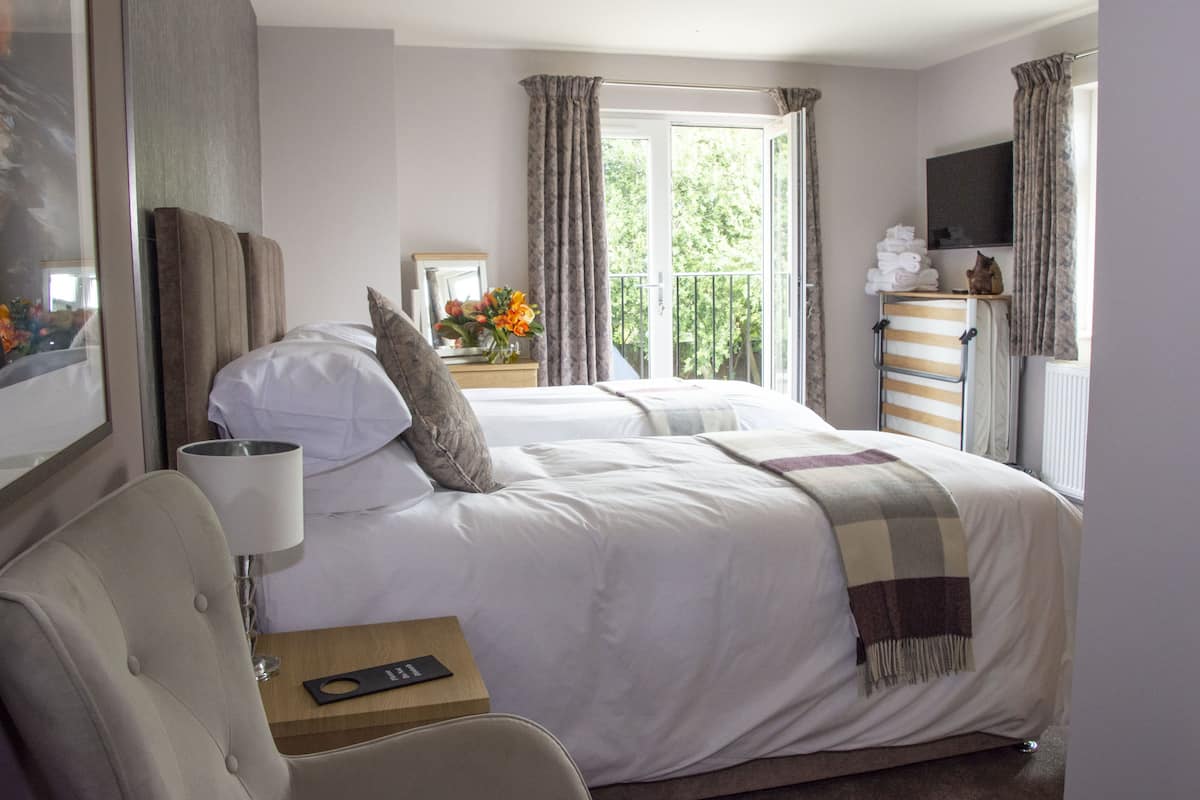 Habitación de hotel con cama, una silla, mesita de noche, lámpara, ventana con cortinas y vista a zonas verdes.