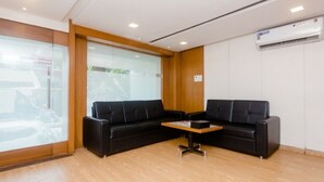 A modern waiting area with two black leather sofas, a wooden coffee table, and a glass wall with blinds. - Hotel Qubestay Airport (Mumbai)