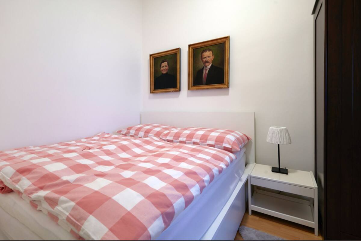 Un dormitorio con una colcha a cuadros, una mesita de noche con lámpara y dos retratos enmarcados en la pared.