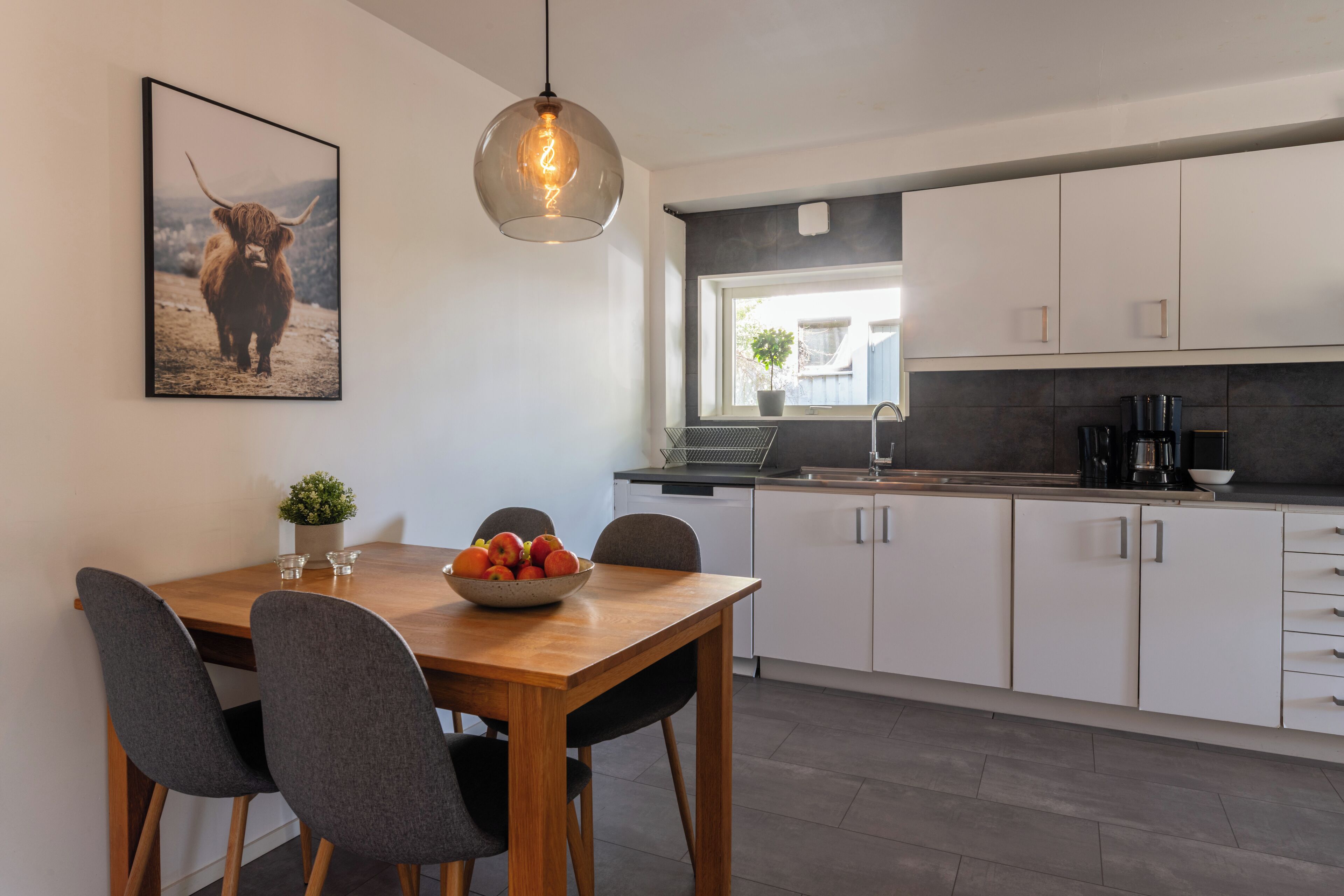A modern kitchen with white cabinets, a wooden dining table, and a hanging light fixture.