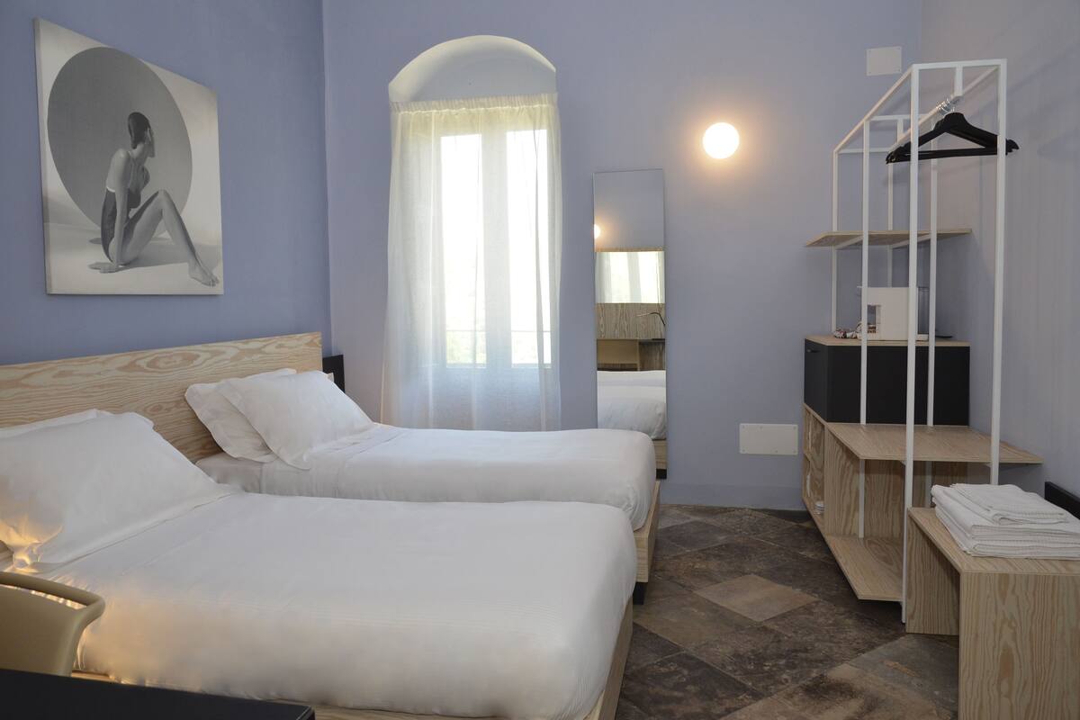 Une chambre d’hôtel avec deux lits, une grande fenêtre avec des rideaux légers, une armoire avec un miroir et un porte-manteau.