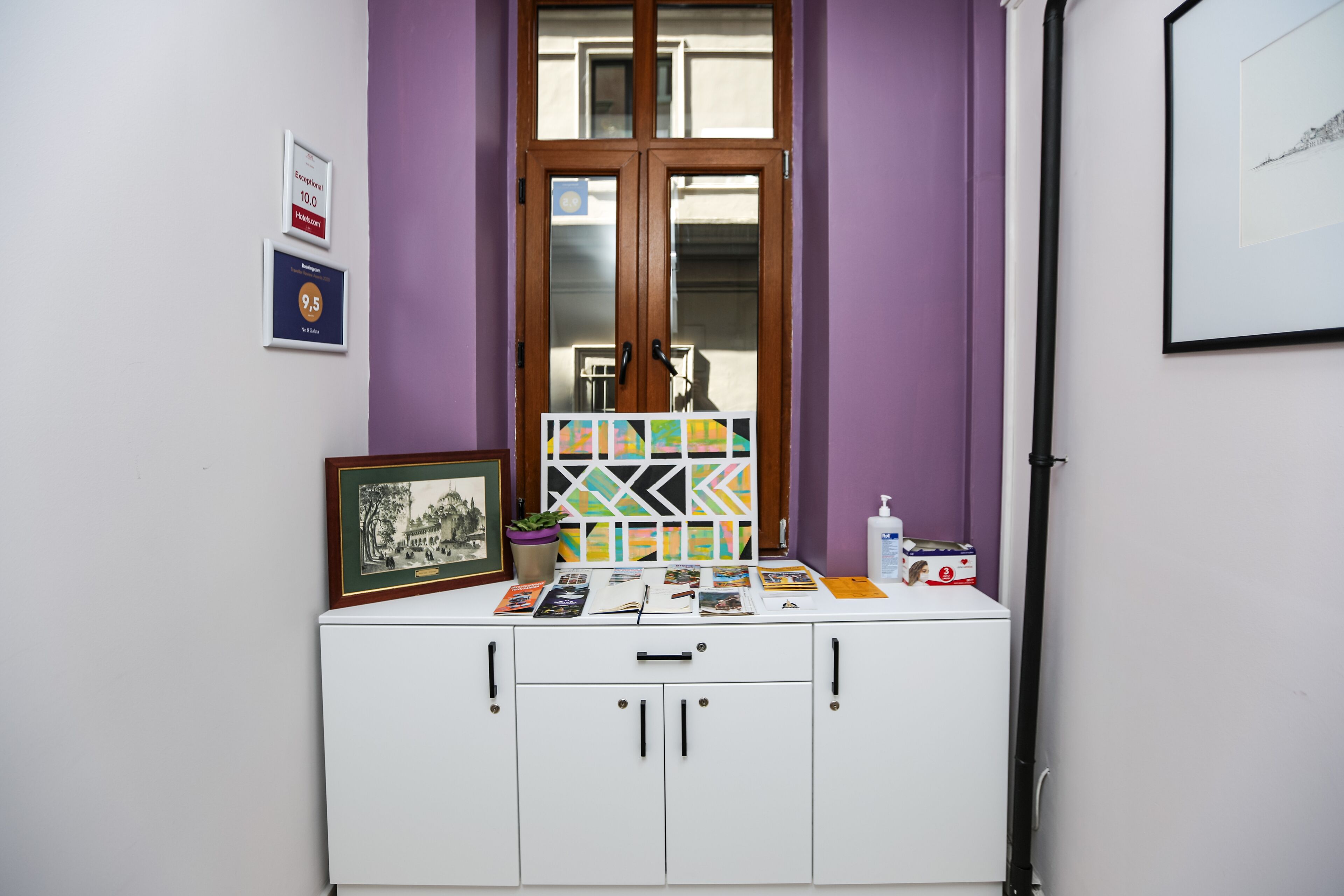 A room with a purple wall, a window with a wooden frame, and a white cabinet with drawers. There are framed certificates on the wall and a small table with various items on top.