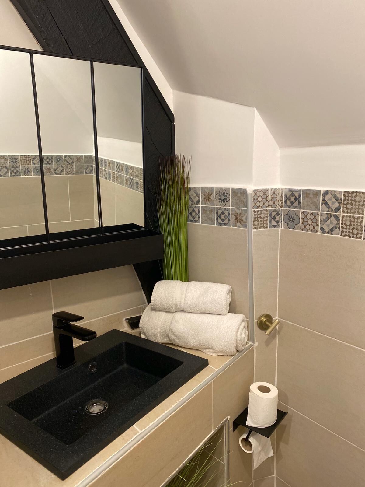 Un baño con un lavabo negro, un espejo, un perchero con toallas, un porta papel higiénico y baldosas decorativas en la pared.