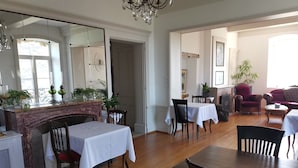 Un comedor con chimenea, mesas y sillas de madera. Se observa un espejo grande sobre la chimenea y un candelabro colgando del techo.