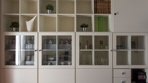 Um armário branco com várias prateleiras e gavetas, contendo utensílios de cozinha, como copos, louças e plantas em vasos.