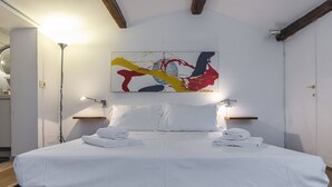 Cama bem arrumada com roupa de cama branca, uma grande pintura abstrata acima e um vigamento de madeira no teto.