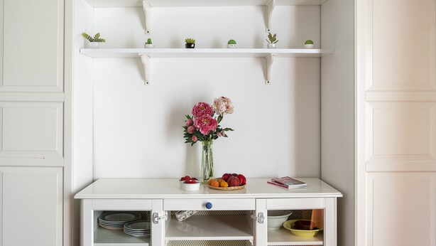 Um armário branco com várias prateleiras, um vaso com flores e uma fruteira.