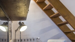 Uma mesa de madeira com uma lâmpada, uma cadeira e livros. Uma escada de madeira é visível ao fundo.