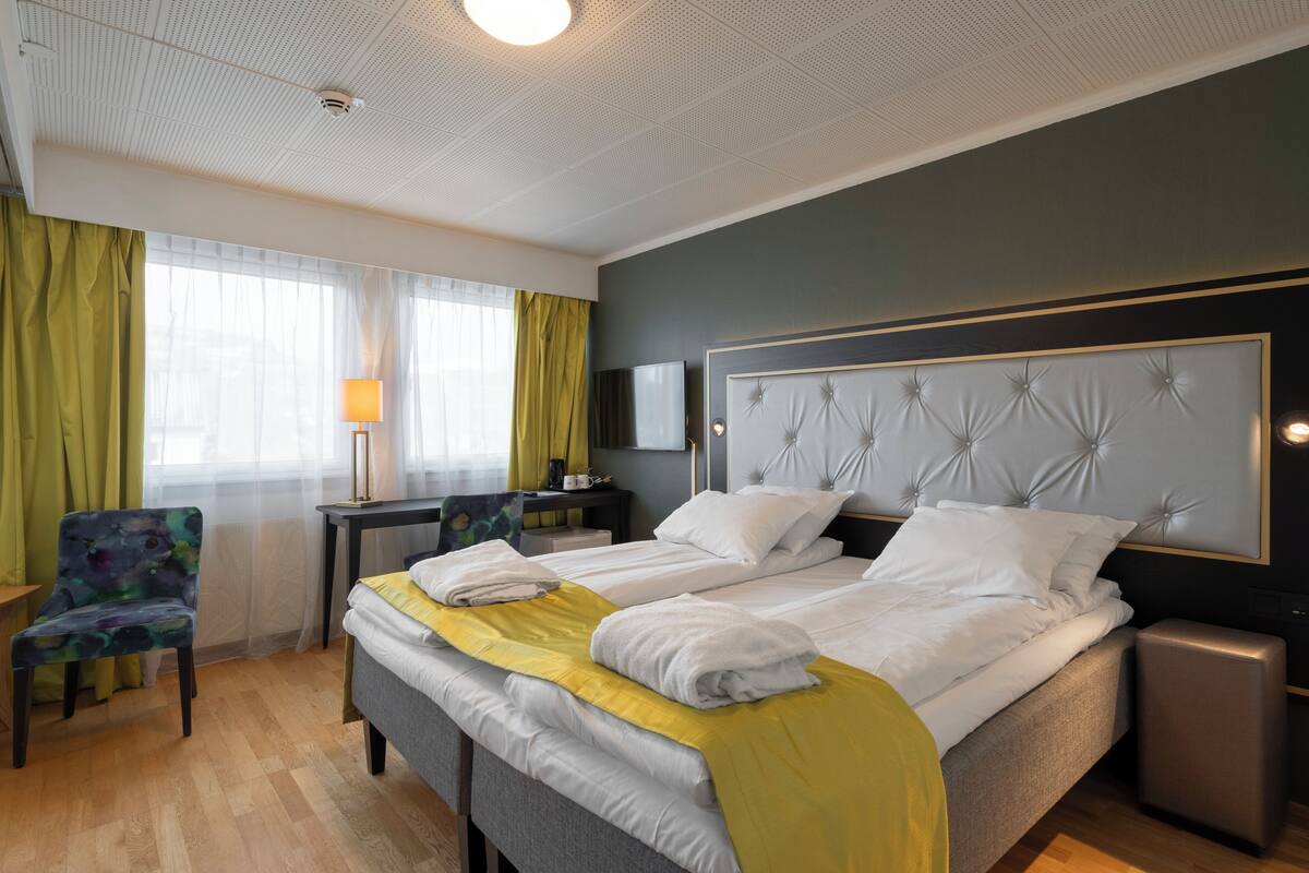 A hotel room with a double bed, a desk, a chair, a bedside table, and a large window with curtains.