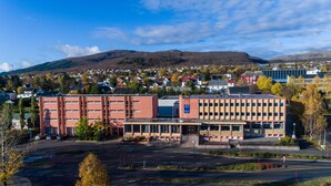 A hotel with a modern design, featuring a central entrance with steps, a parking lot, and a view of a town and mountains in the background.
