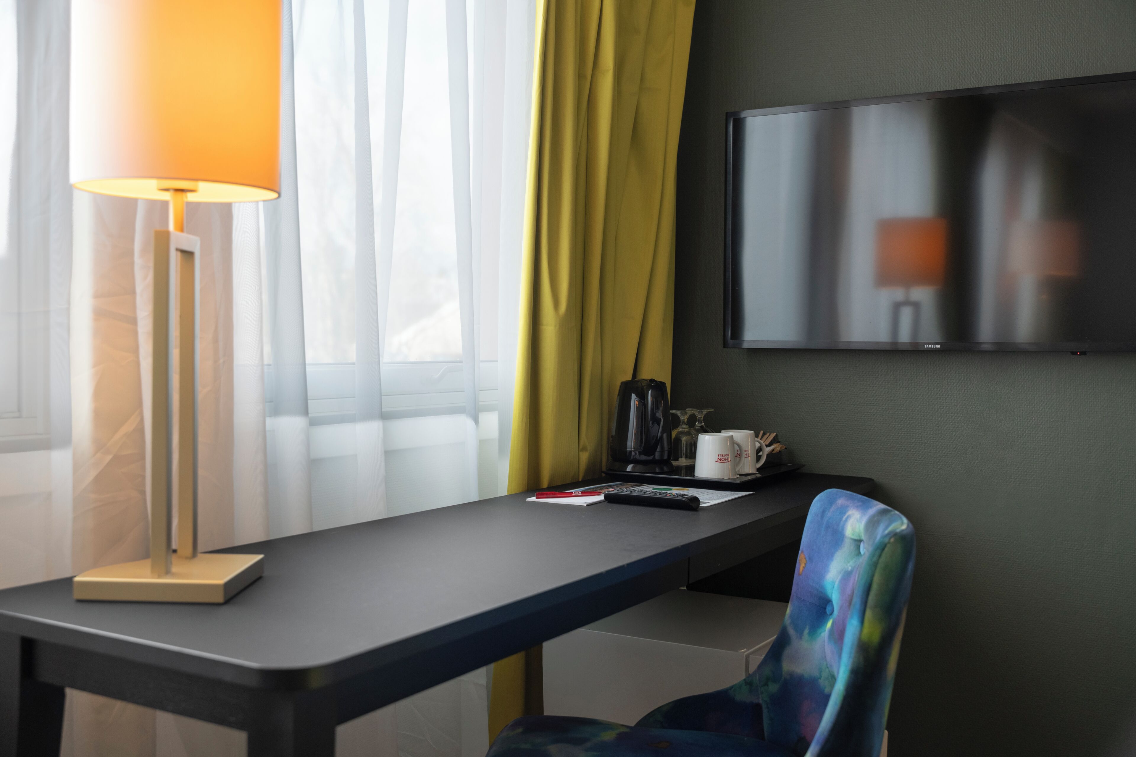 A hotel room with a desk, a lamp, a chair, a flat-screen TV, and a window with curtains.