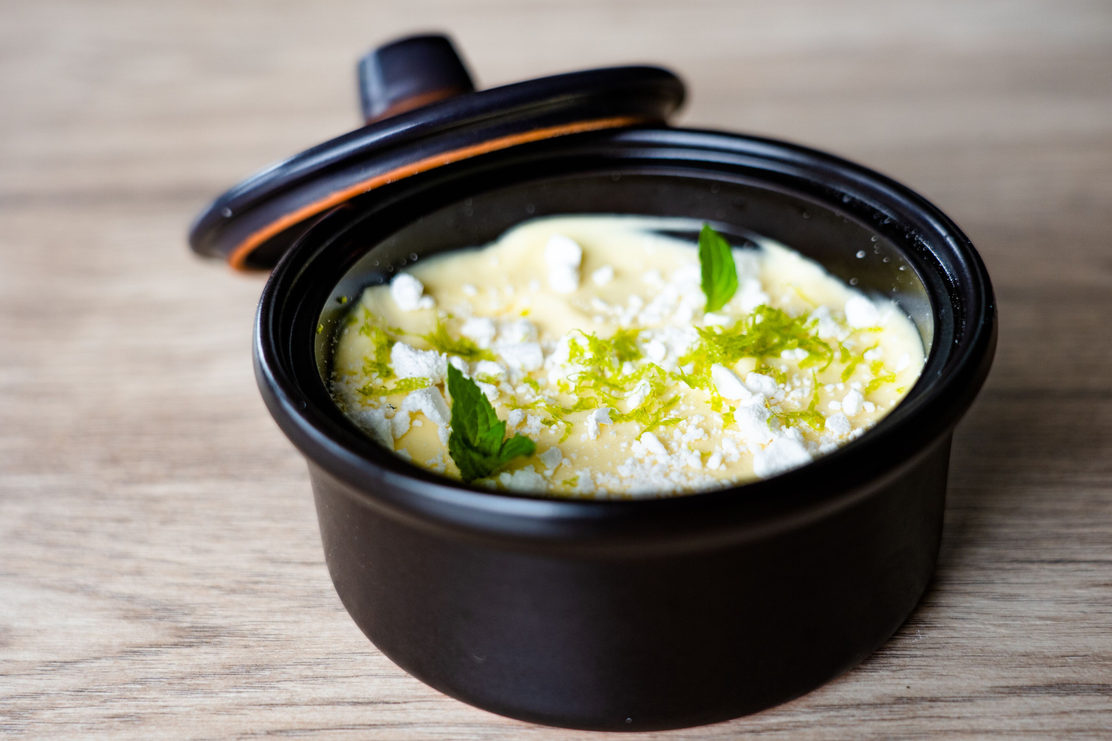 A black ceramic bowl with a lid, containing a creamy soup garnished with herbs, on a wooden surface.