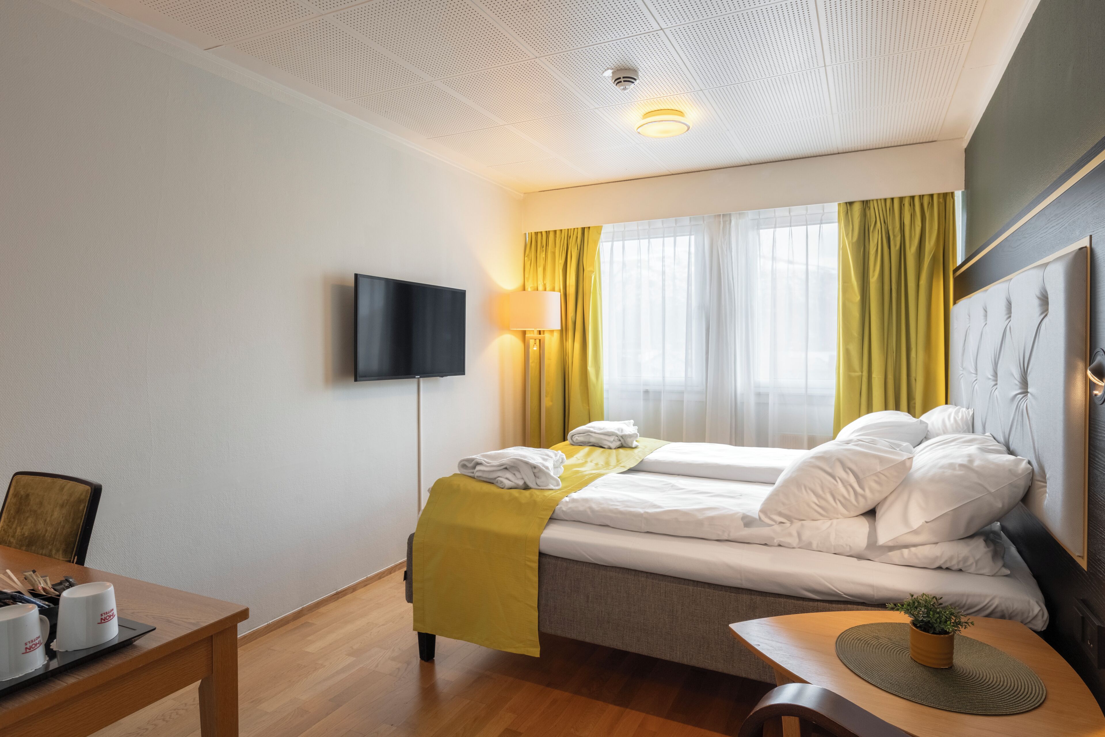 A hotel room with a bed, a desk with a chair, a flat-screen TV, a lamp, and a window with curtains.