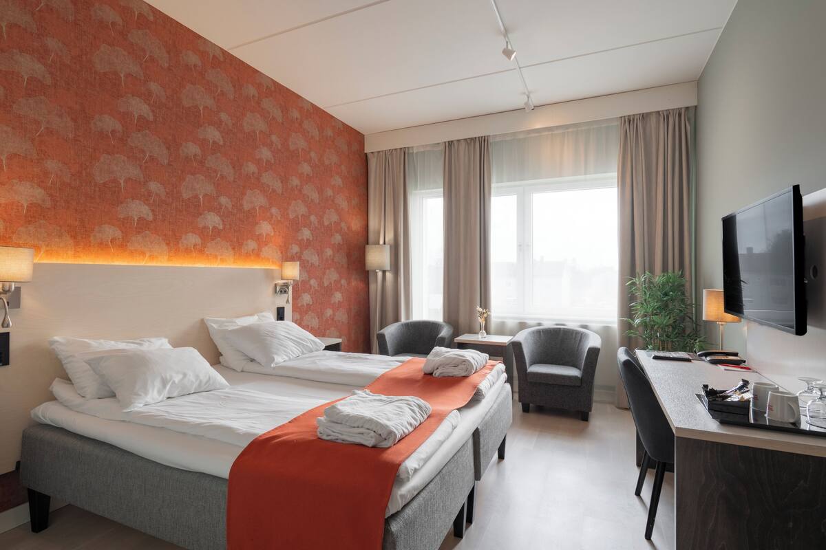 A hotel room with a bed, two chairs, a desk, a TV, and a window with curtains.