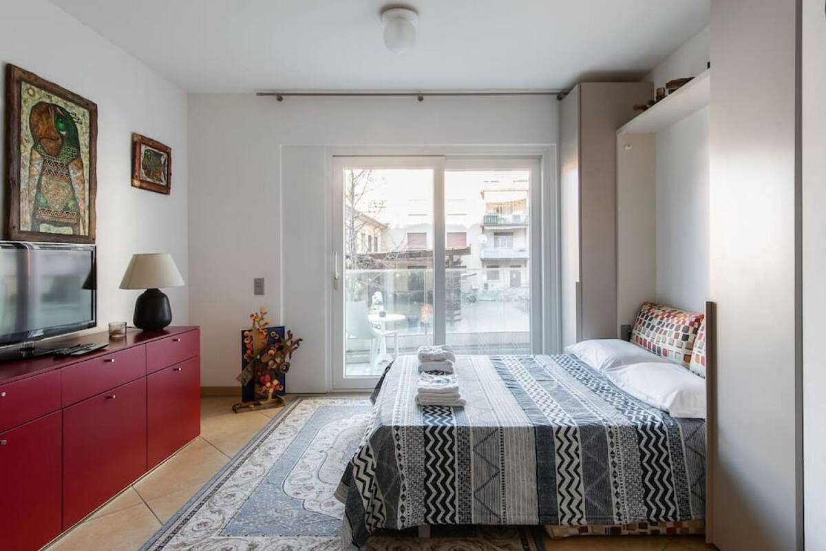 Una camera da letto con un letto, una TV su un mobiletto rosso e una finestra vista edifici.