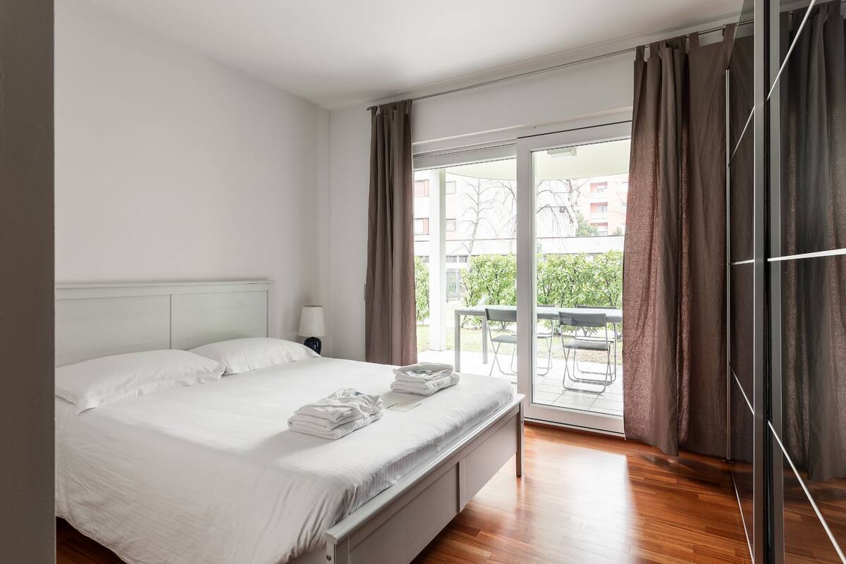 Camera d'albergo con un letto, un comodino, una lampada e vista sull'esterno tramite una porta scorrevole in vetro.