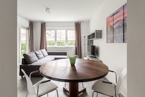 Ein modernes Wohnzimmer mit einem runden Holztisch, zwei weißen Stühlen, einem grauen Sofa und einem großen Fenster mit Vorhängen.