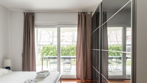 A modern hotel room with a large bed, a view of the outdoor patio, and a sliding glass door. - Prestige Du Lac Apartments (Lugano)