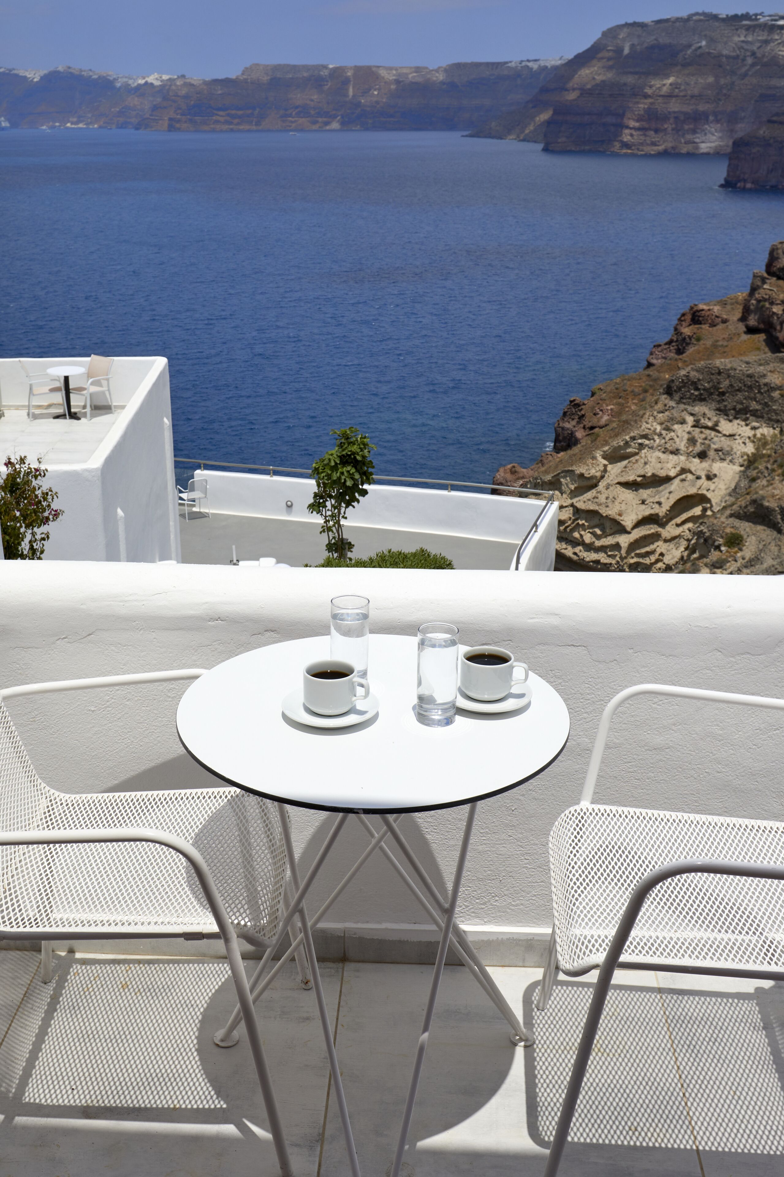 Un espacio exterior con asientos blancos, una mesa y sillas, con vista a un cuerpo de agua y acantilados rocosos.