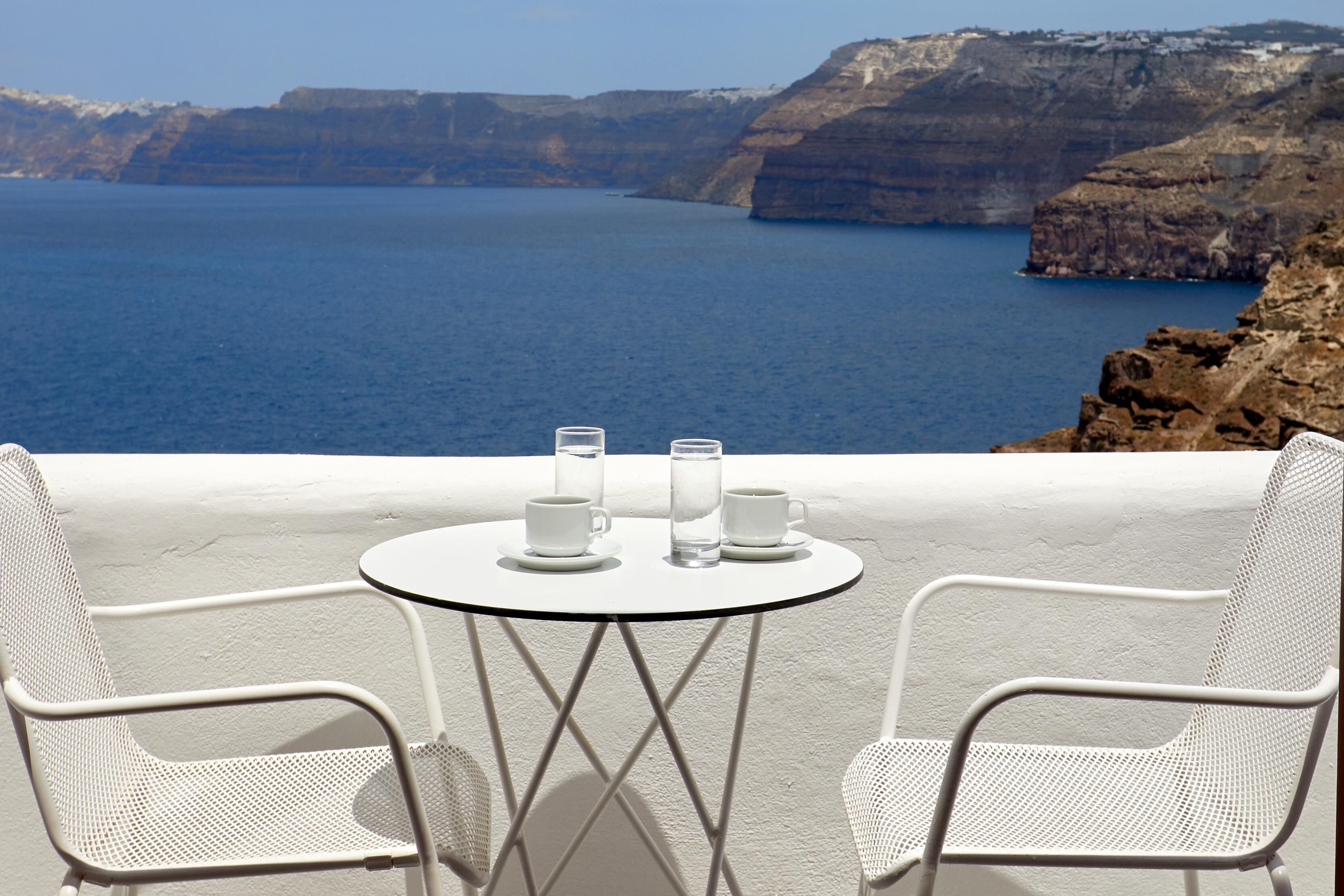 A white metal table with two chairs and a glass of water, overlooking a coastal cliff and sea.