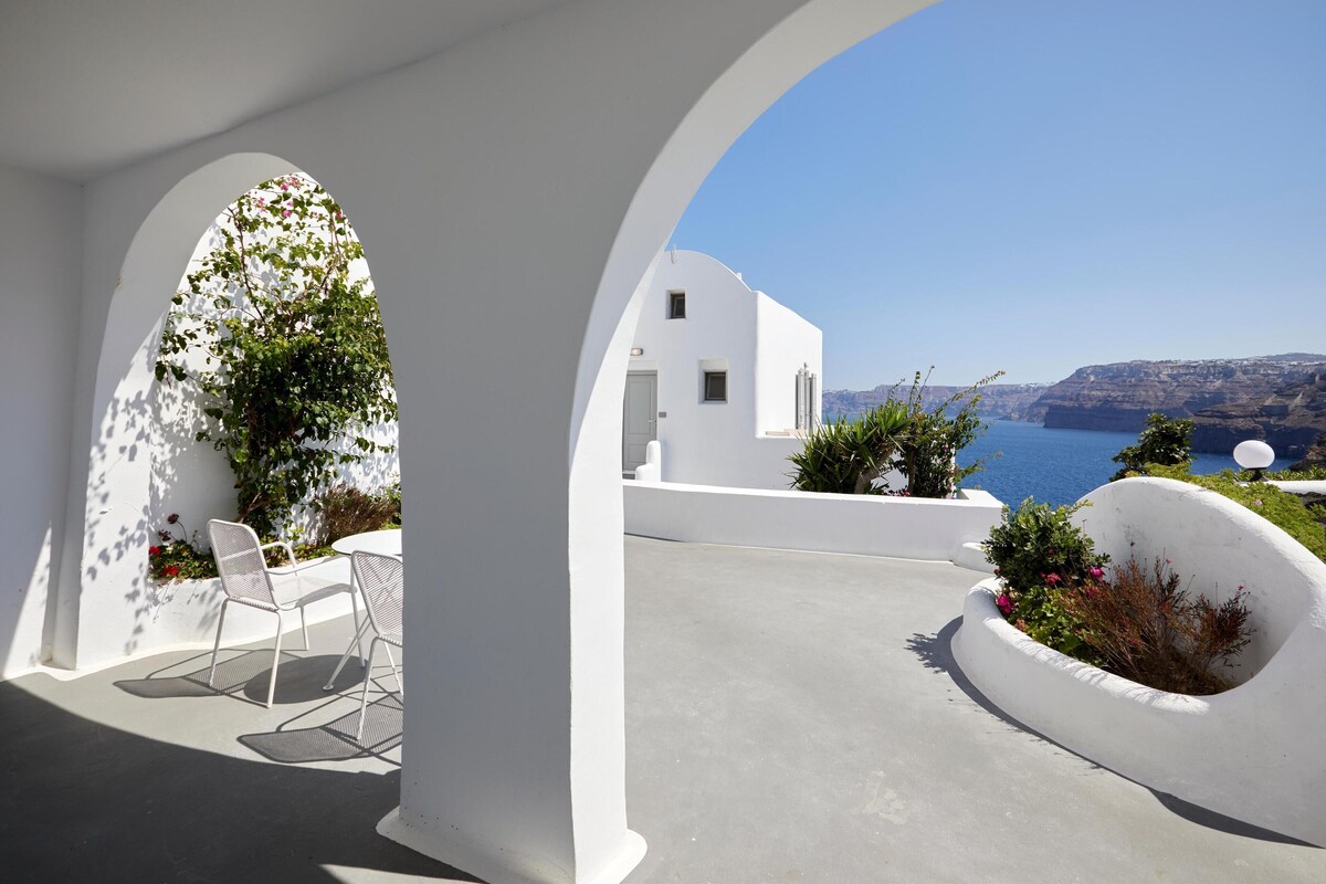 A white-walled outdoor area with a table and chairs, a white planter with plants, and a view of the sea and cliffs.