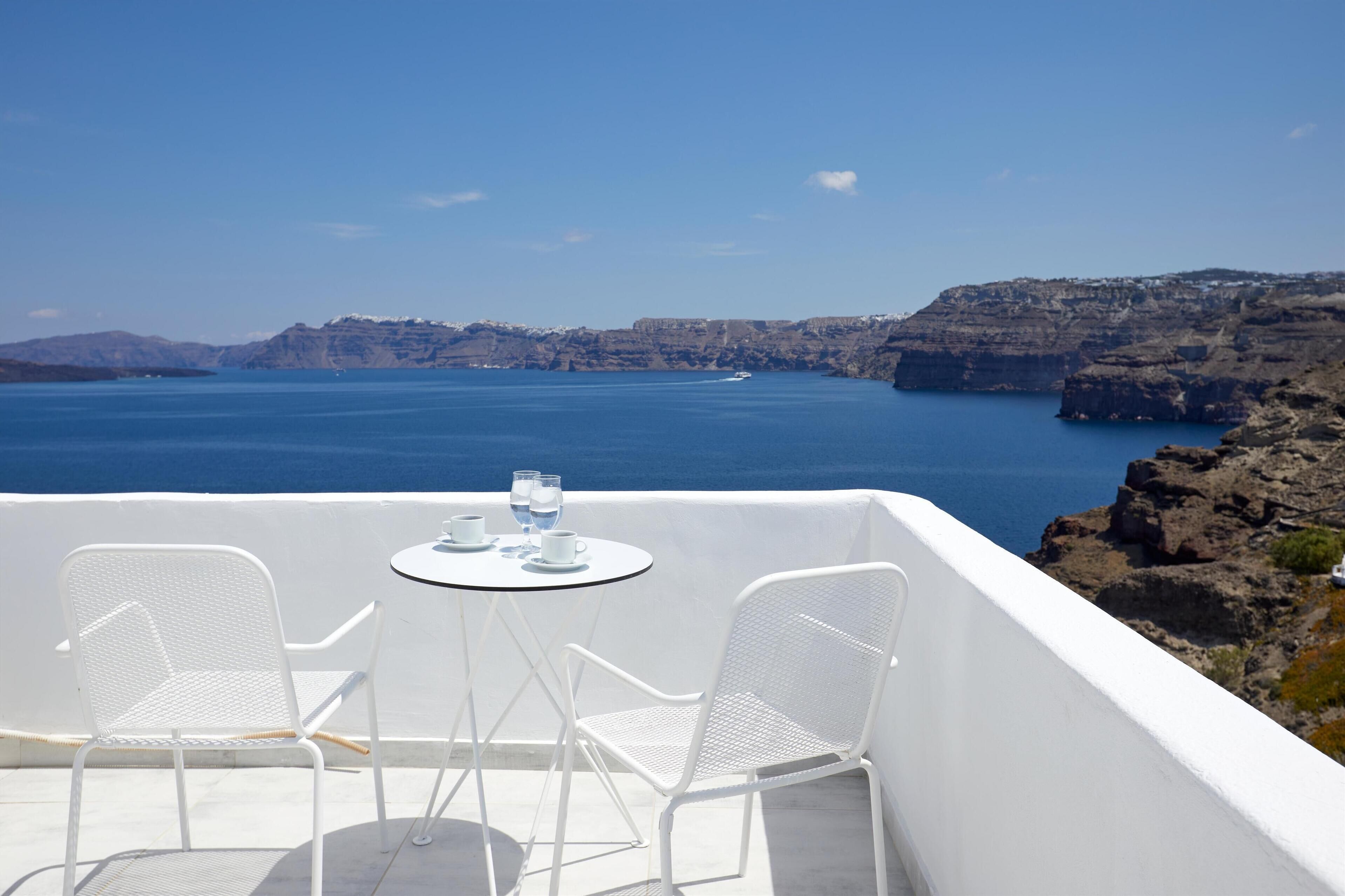 A terrace with white metal chairs and a small round table, overlooking a large body of water and rocky cliffs.