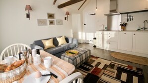 A cozy living room with a sofa, a dining table with breakfast items, and a kitchen with white cabinets and a hanging knife rack. - Fazeboons Barn (Woodbridge)