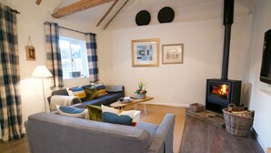 A cozy living room with a sofa, a table, a fireplace, and a window with curtains. - Fazeboons Barn (Woodbridge)