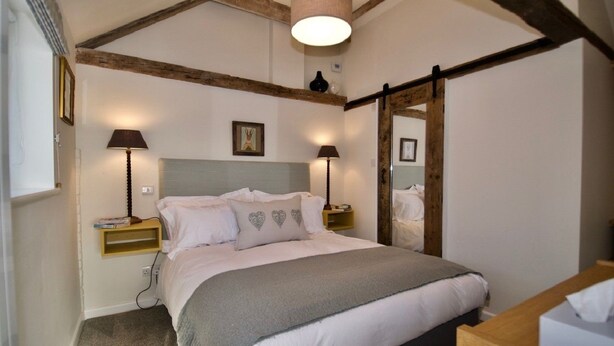 A bedroom with a bed, bedside lamps, a mirror, and a wooden ceiling beam. - Fazeboons Barn (Woodbridge)
