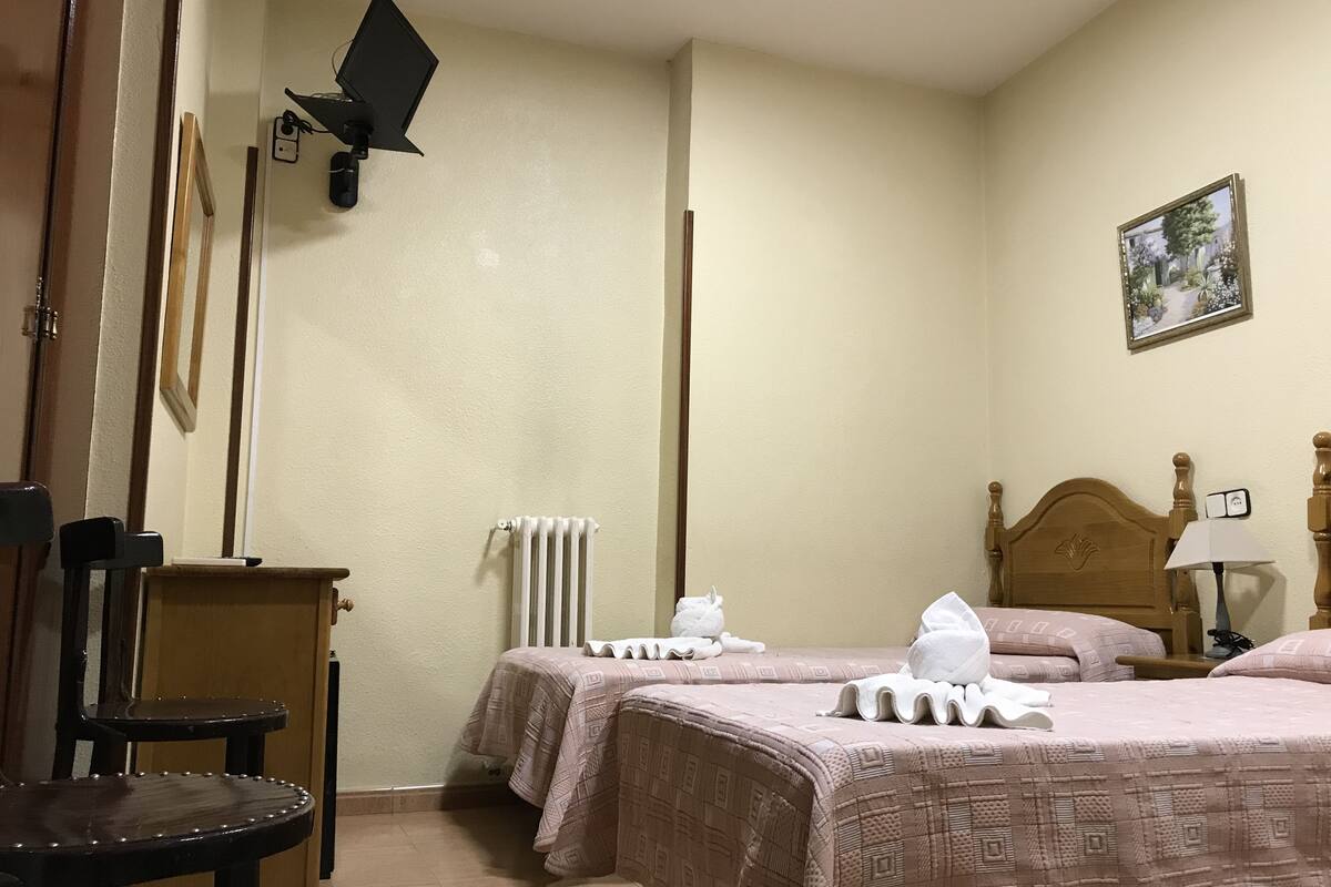 A hotel room with two beds, a wooden chair, a radiator, and a painting on the wall.