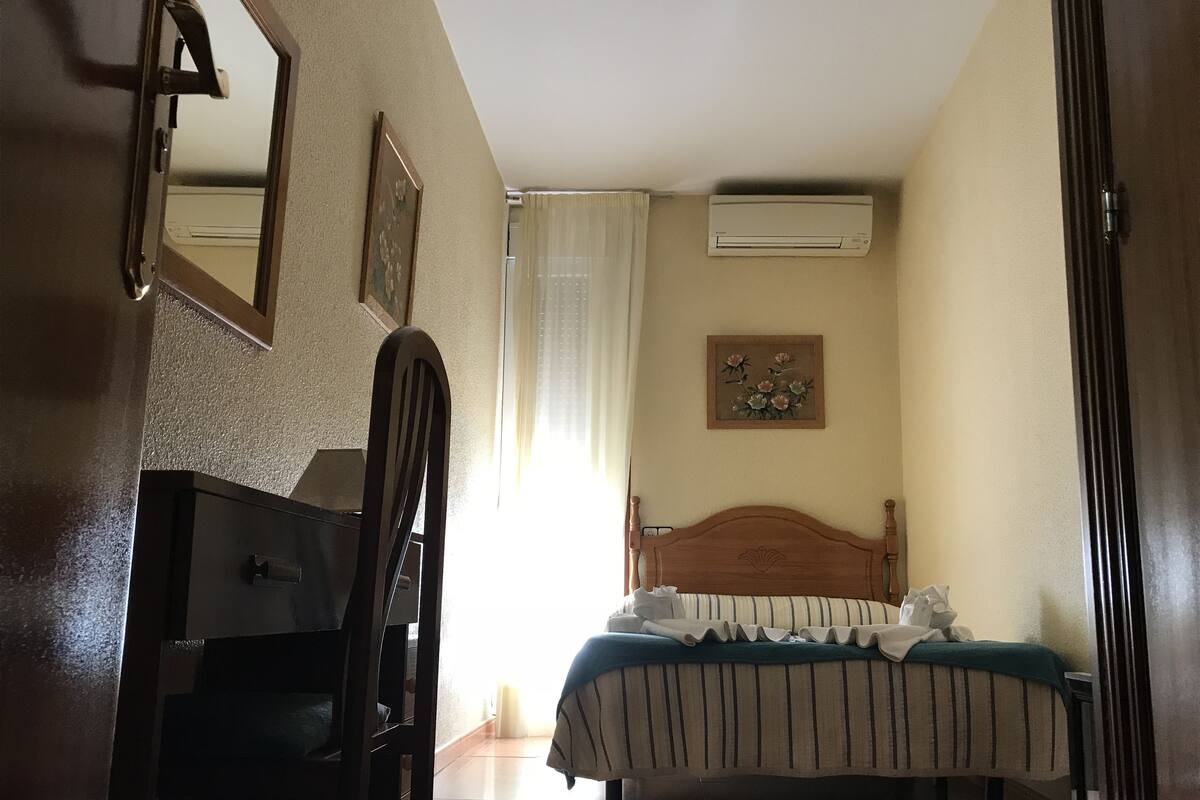 A bedroom with a bed, a wooden chair, a dresser, a mirror, and an air conditioning unit.