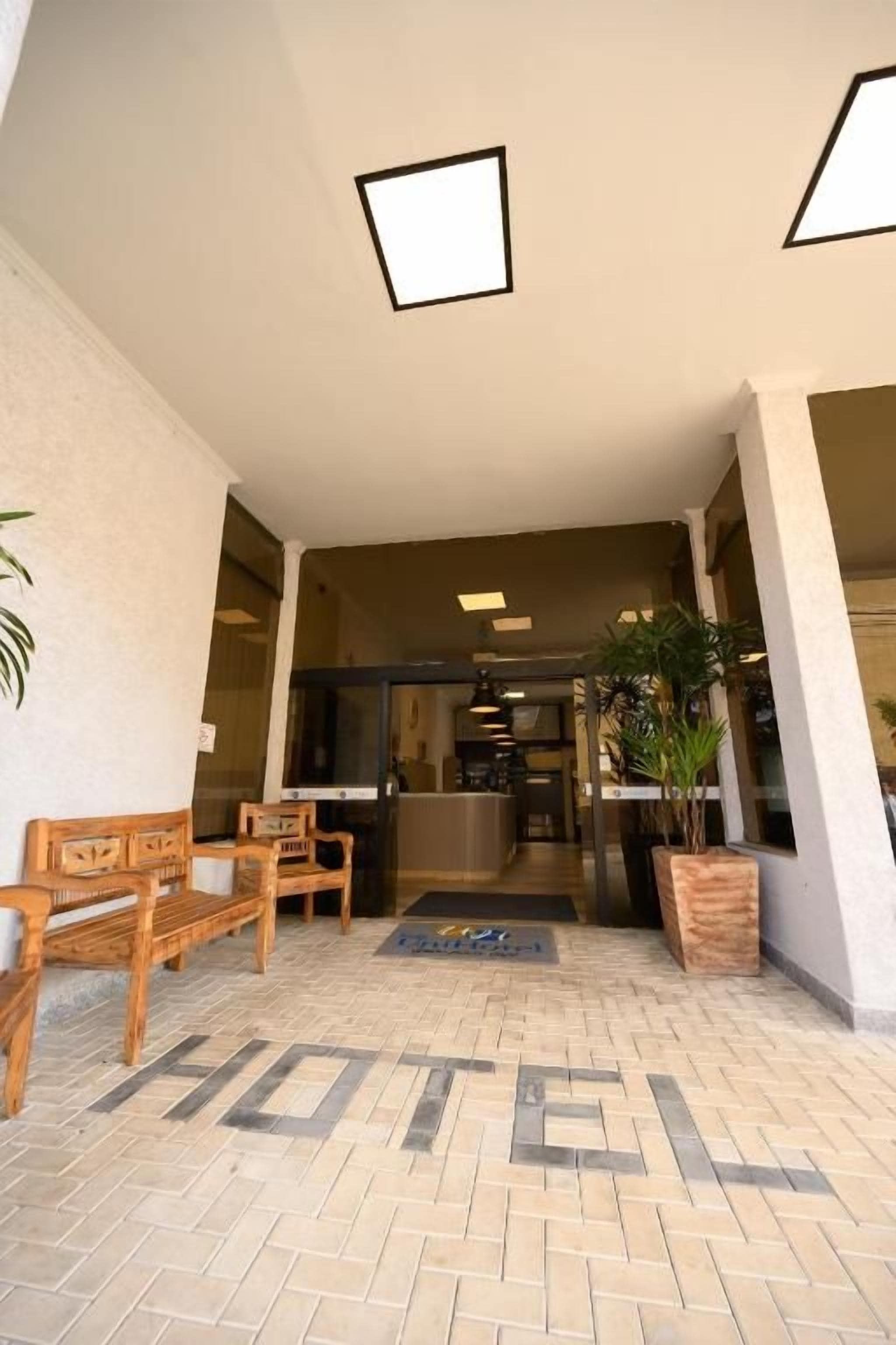 A hotel lobby with a reception desk, wooden benches, a potted plant, and a tiled floor.
