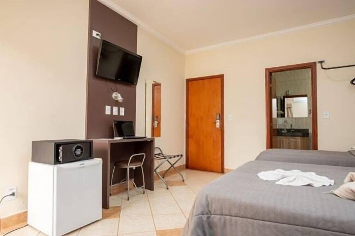 A hotel room with a bed, a desk with a chair, a television mounted on the wall, and a small refrigerator.