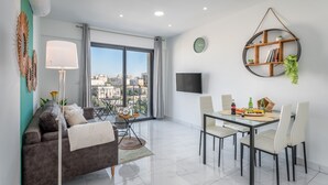 A modern living room with a sofa, dining table, chairs, a TV, and a large window with a city view. - Even Israel Apartments (Jerusalem)