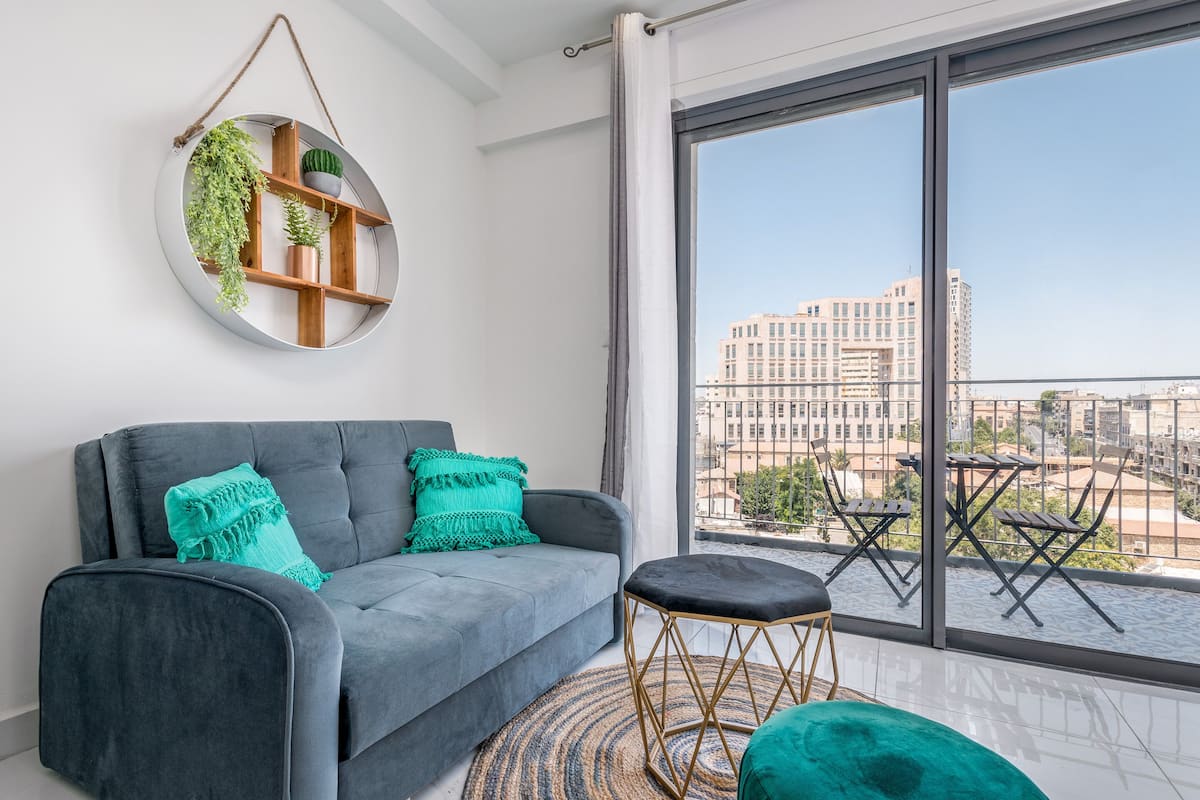 Ein modernes Wohnzimmer mit einem grauen Sofa, türkisfarbenen Kissen, einem runden Spiegel mit Pflanzen und Blick auf Gebäude durch große Fenster.