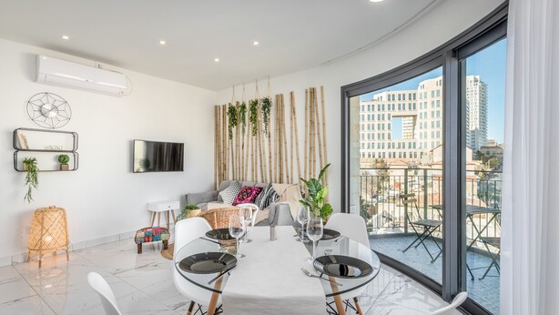 A modern dining area with a white table set for six, a sofa with cushions, a TV mounted on the wall, and a view of buildings through large windows. - Even Israel Apartments (Jerusalem)