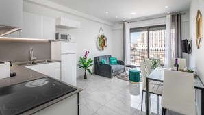 A modern kitchen with a built-in stove, a dining area with a table and chairs, a gray sofa with teal cushions, and a large window offering a city view. - Even Israel Apartments (Jerusalem)