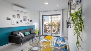 A modern living room with a black sofa, a glass dining table, and a balcony with a view of buildings. - Even Israel Apartments (Jerusalem)