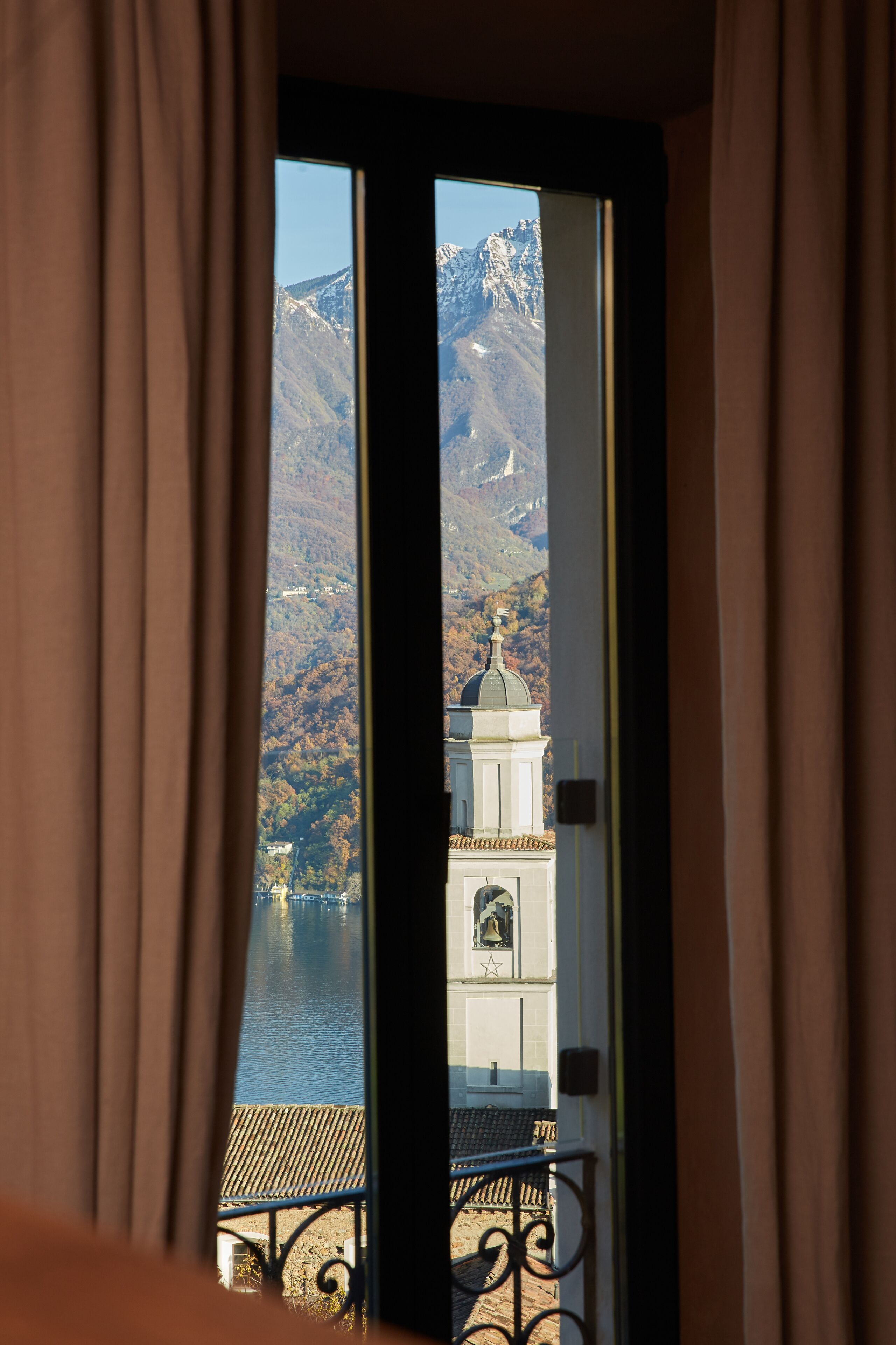 Blick durch ein Fenster mit Vorhängen, der eine Berglandschaft, ein Gewässer und ein Gebäude mit Kuppel und Glockenturm zeigt.