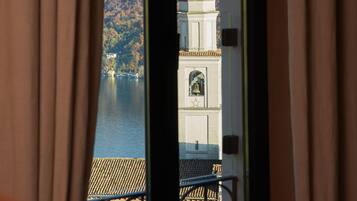 Blick durch ein Fenster mit Vorhängen, der eine Berglandschaft, ein Gewässer und ein Gebäude mit Kuppel und Glockenturm zeigt.