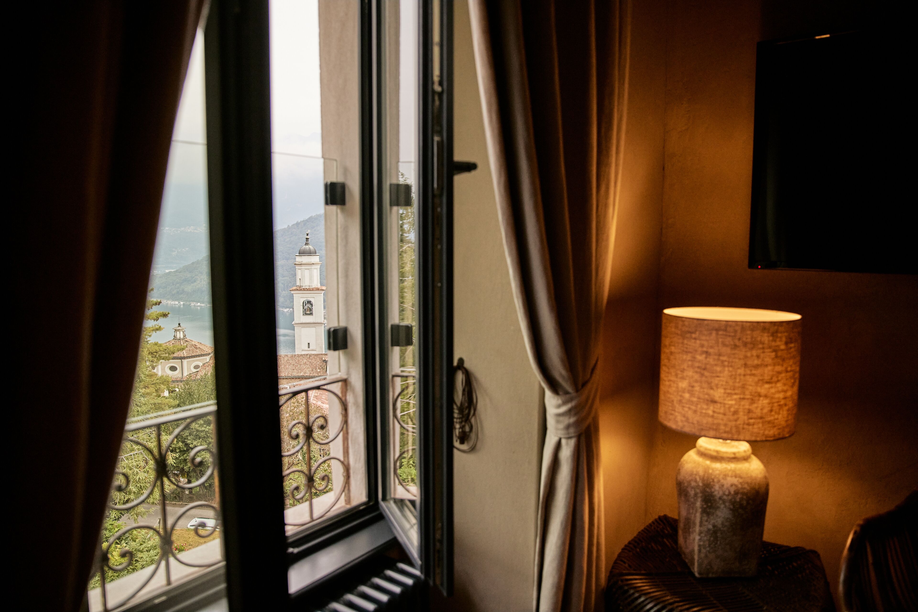 A room with a view of a building through a window, a lamp on a side table, and a curtain partially drawn.