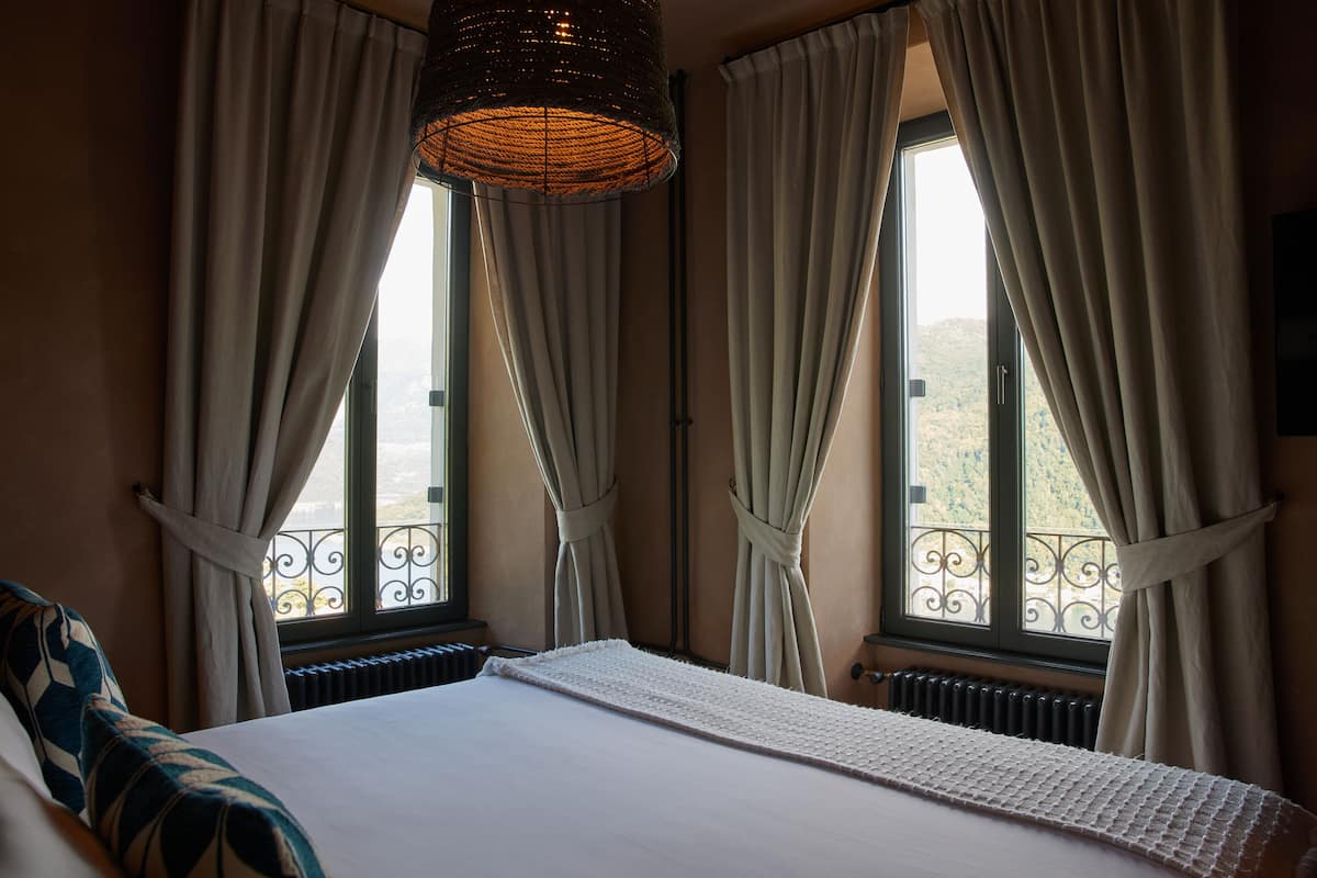 Quarto com uma cama grande, duas janelas com cortinas, um radiador e vista para uma varanda.