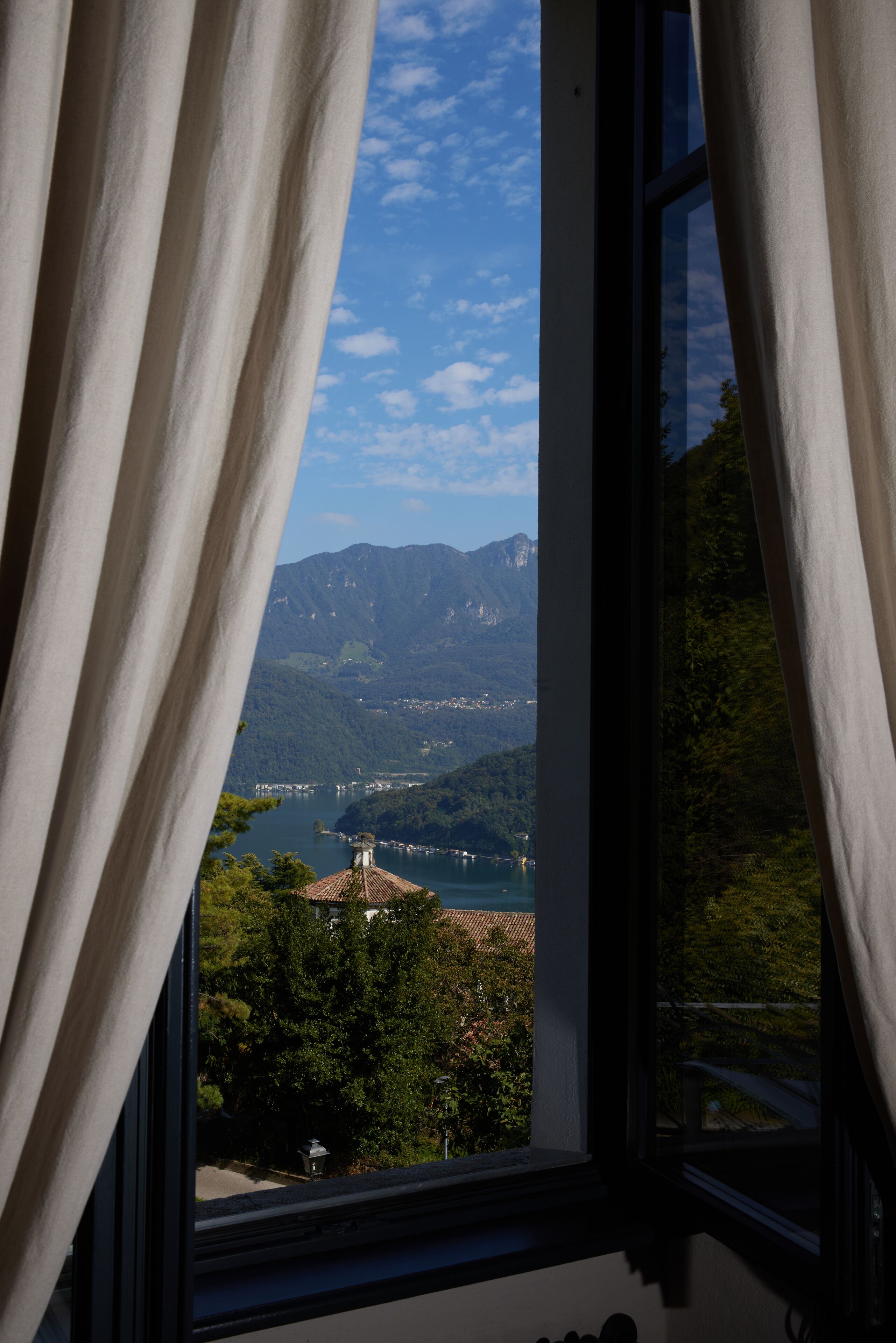 Vista a través de una ventana con cortinas, mostrando un paisaje montañoso con un lago y edificios a lo lejos.