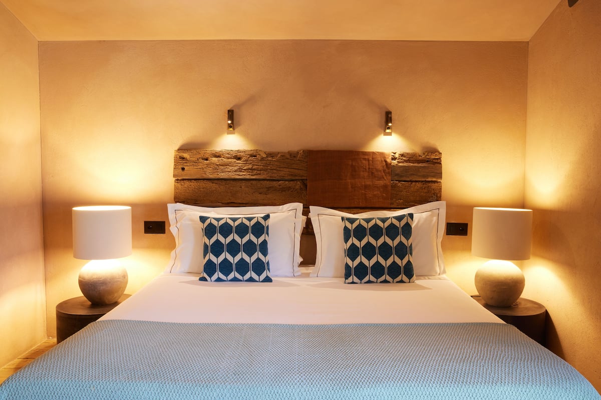 Quarto de hotel com uma cama grande, duas luminárias fixadas na parede e uma cabeceira de madeira rústica.