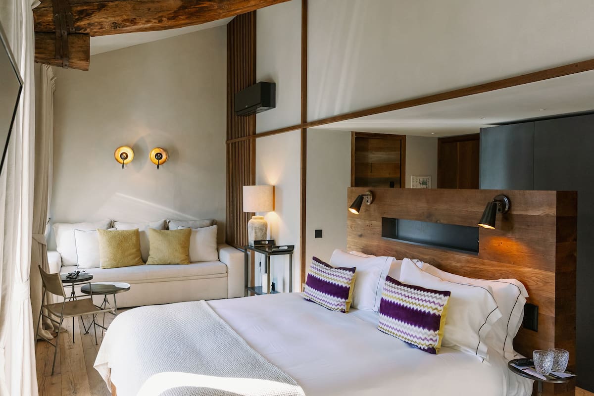 Quarto de hotel moderno com uma cama grande, um sofá, uma mesinha e uma TV fixada na parede.