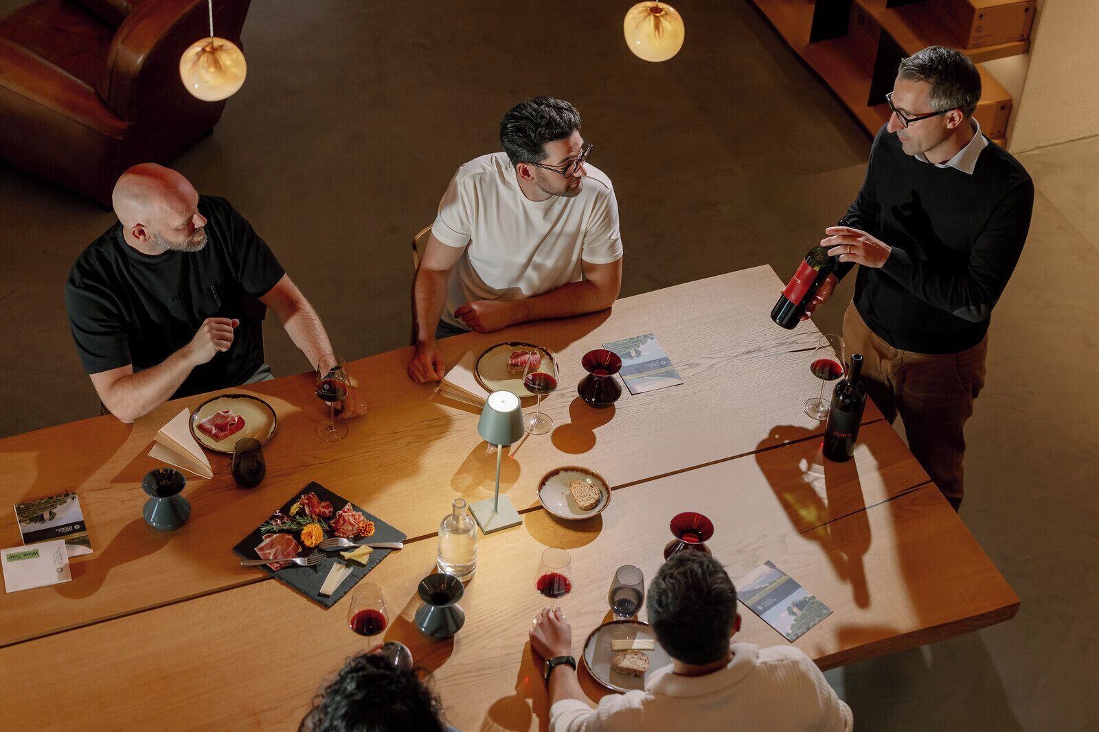 Un grupo de personas sentadas alrededor de una mesa de madera, con copas de vino, platos de comida y una vela.